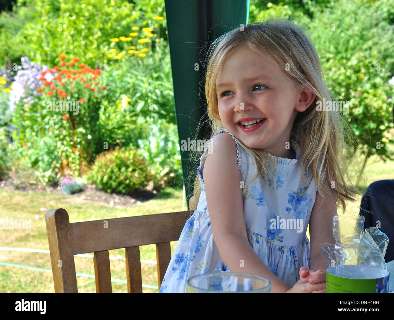 portrait of little blonde girl in blue and white floral dress smiling and giggling seated at a table outside in a garden Stock Photo