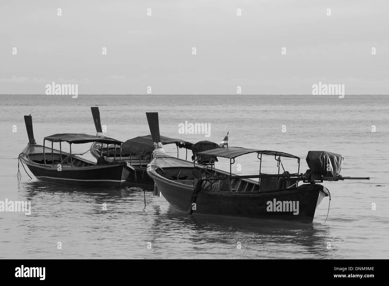 A black and white photograph of some longtail boats in Thailand against a backdrop of a calm sea and horizon. Stock Photo