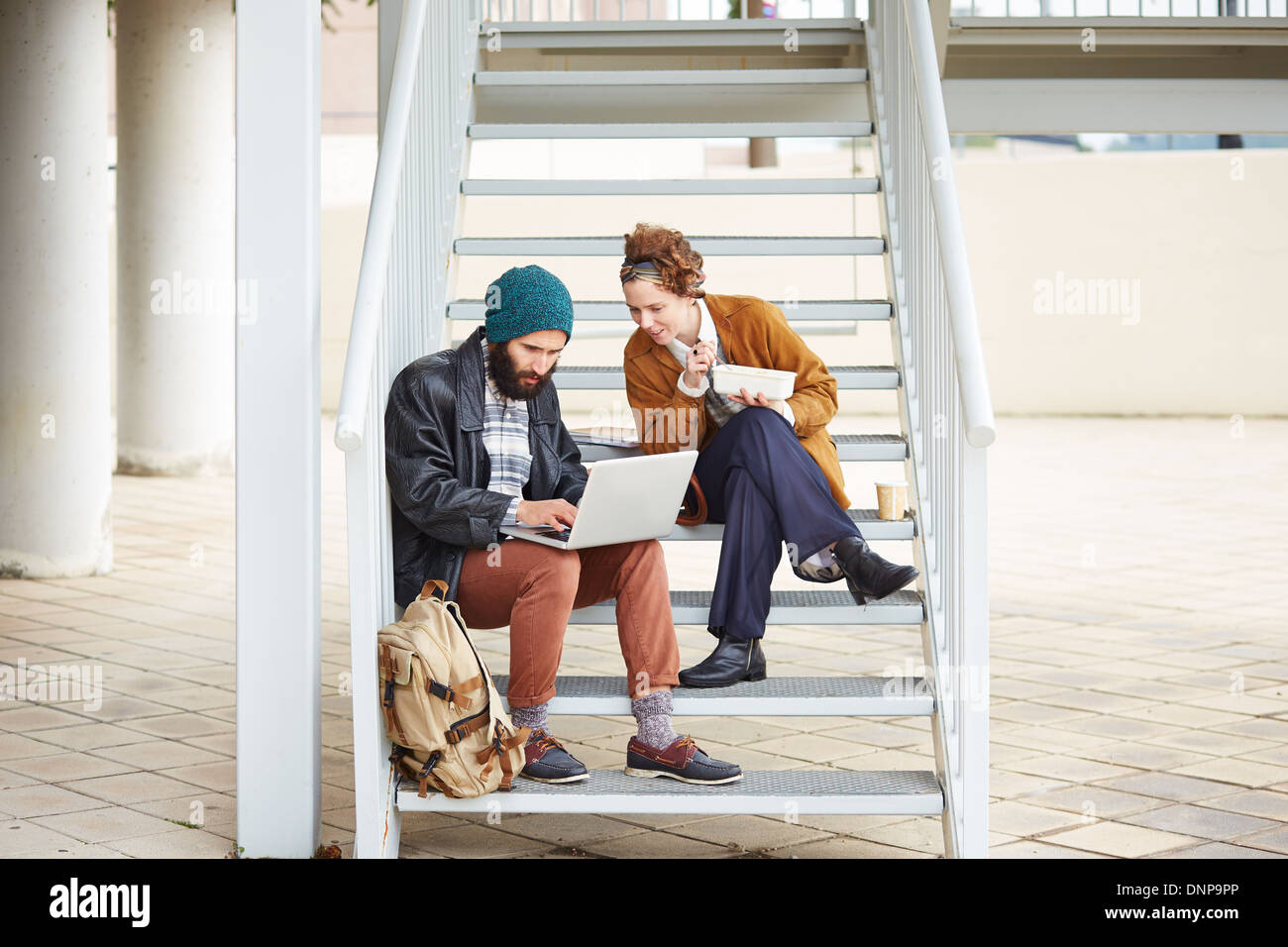 Hipster couple using computer and eating lunch sitting in stairs at university campus Stock Photo