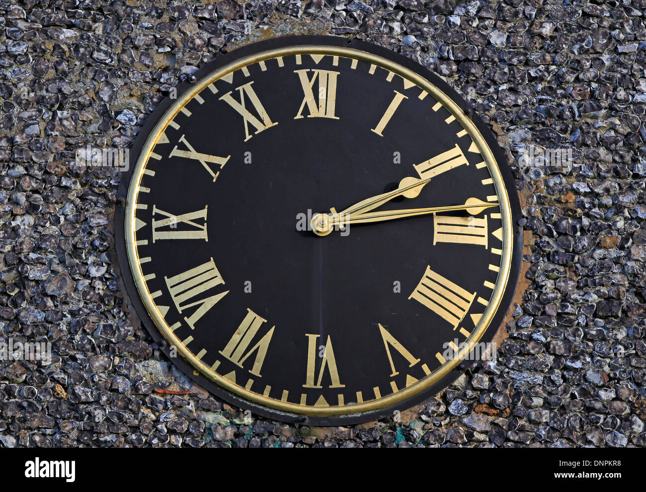 Circular black clock with gold roman numerals attached to a flint wall