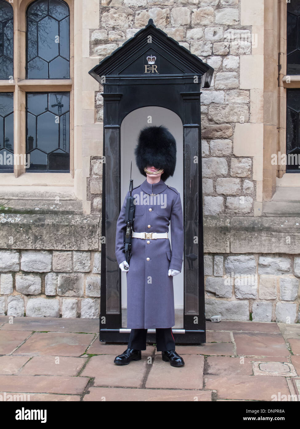 Queen's Guard soldier wearing a greatcoat and busby bearskin hat, standing to attention at a sentry box in the Tower of London Stock Photo