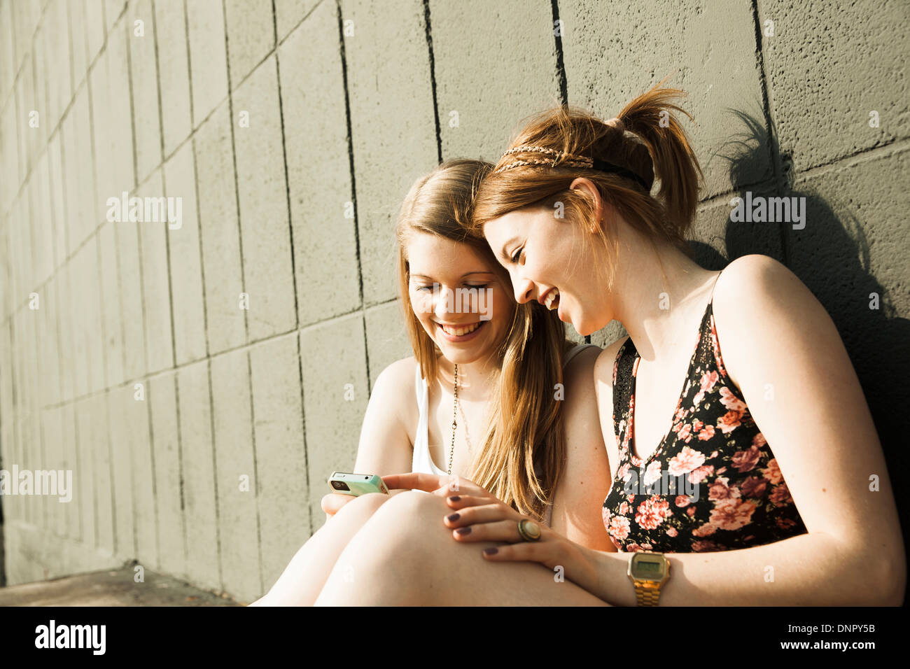 Young women sitting and leaning against wall, looking at smart phone together Stock Photo