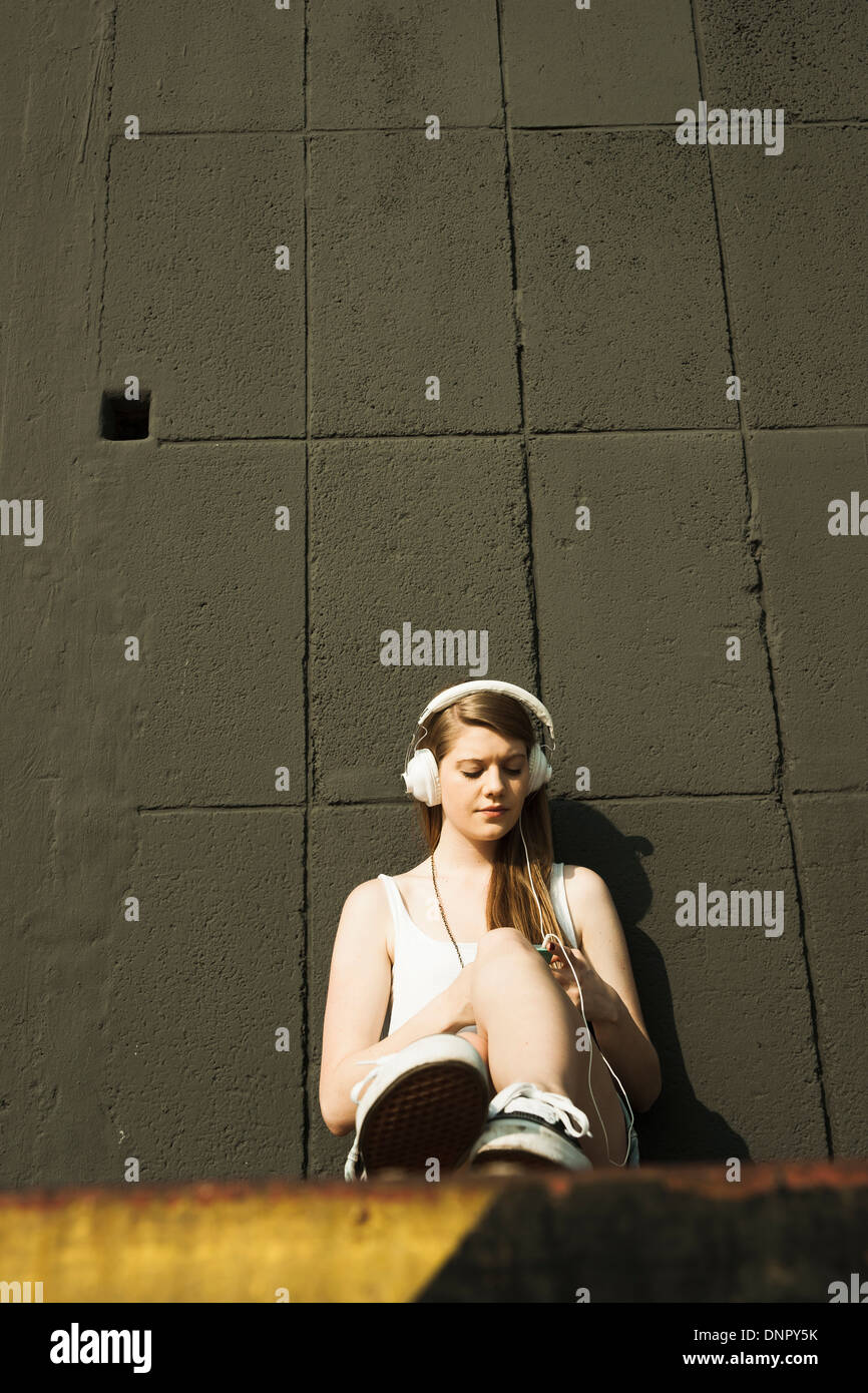 Young woman sitting on ground, leaning against cement wall, listening to MP3 player Stock Photo
