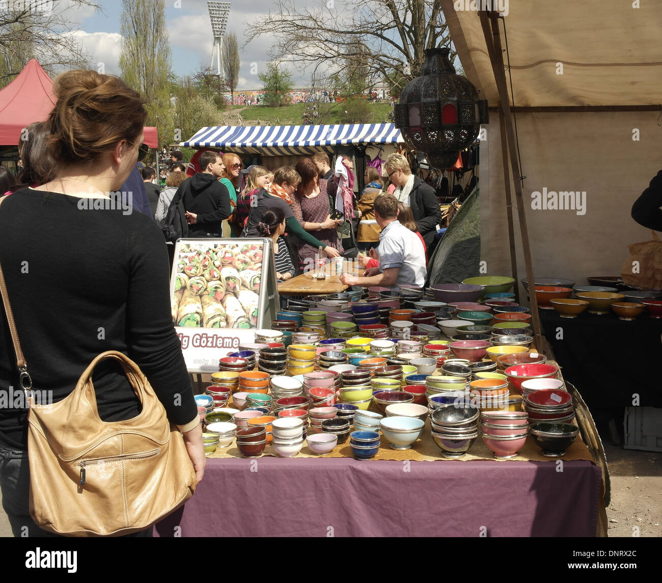 Sunny view young woman standing market stall table with colourful pots, towards people walkiing, Mauerpark Flea Market, Berlin Stock Photo