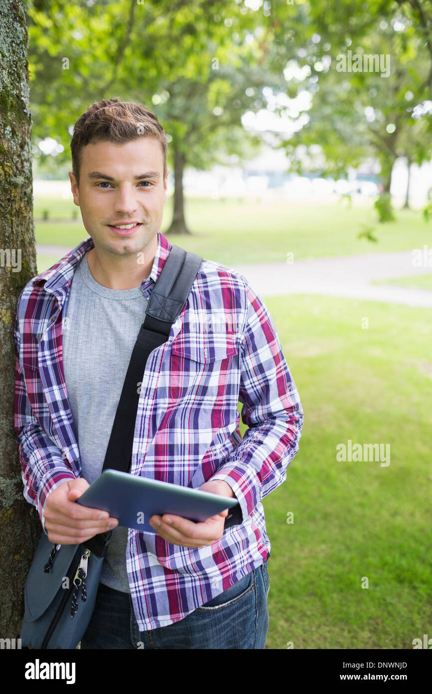 Handsome student leaning on tree looking at camera holding tablet Stock Photo