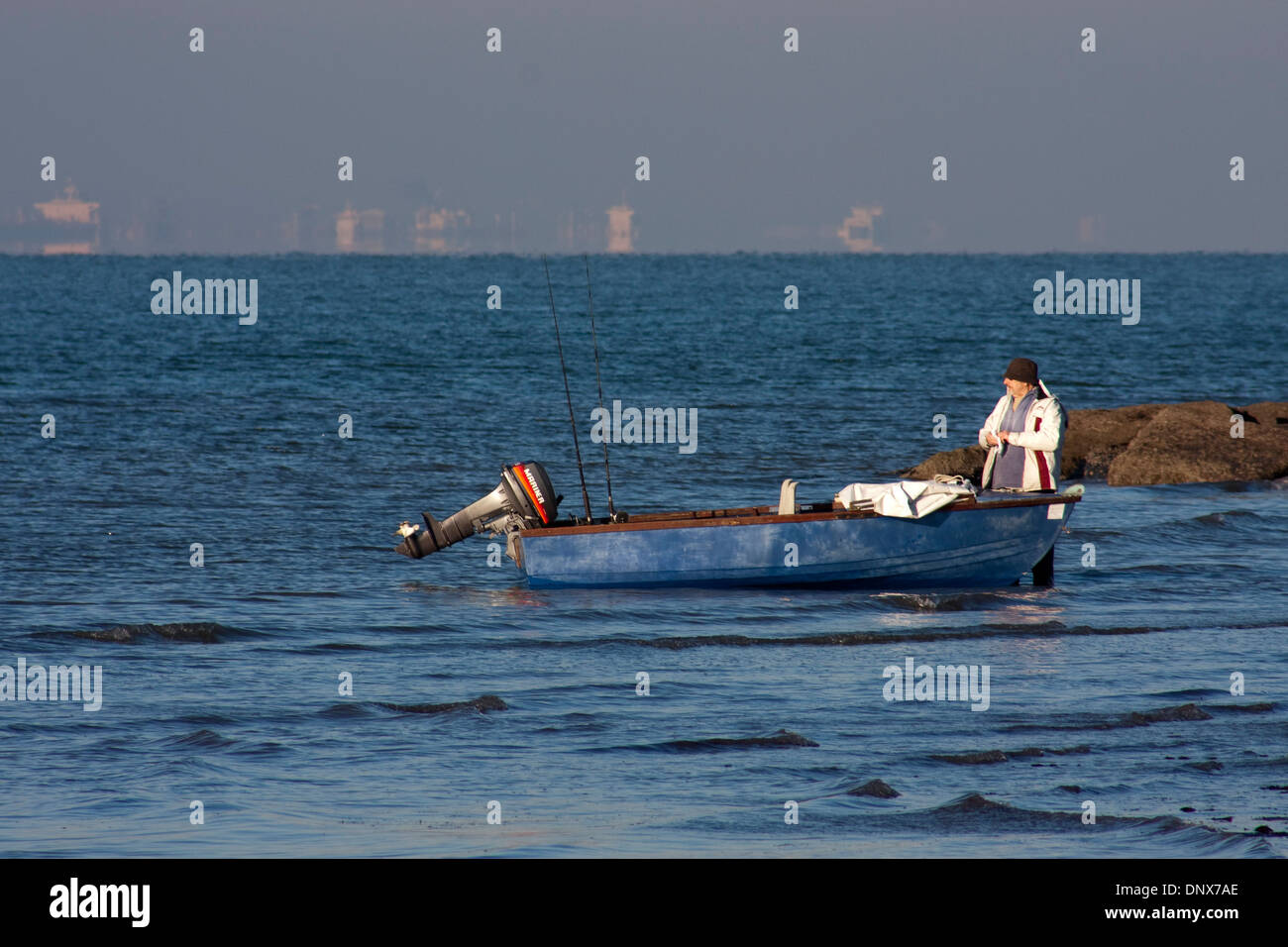 A middle aged man stands in blue water with a fishing dinghy Stock Photo