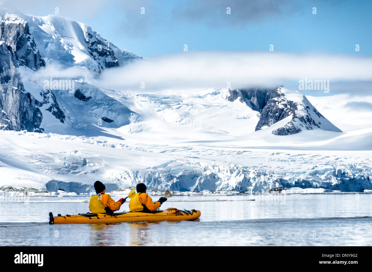 Kayakers Paddle Petermann Island Antarctica // PETERMANN ISLAND, Antarctica — Kayakers paddle through glassy, mirror-like waters along the scenic mountainous shoreline near Petermann Island on the western side of the Antarctic Peninsula. The calm conditions offer perfect reflections of the snow-capped peaks, creating a stunning symmetry between sky and sea in this pristine polar landscape. This area is known for its breathtaking views and diverse wildlife, including penguins and seals, which thrive in the harsh Antarctic environment. Stock Photo