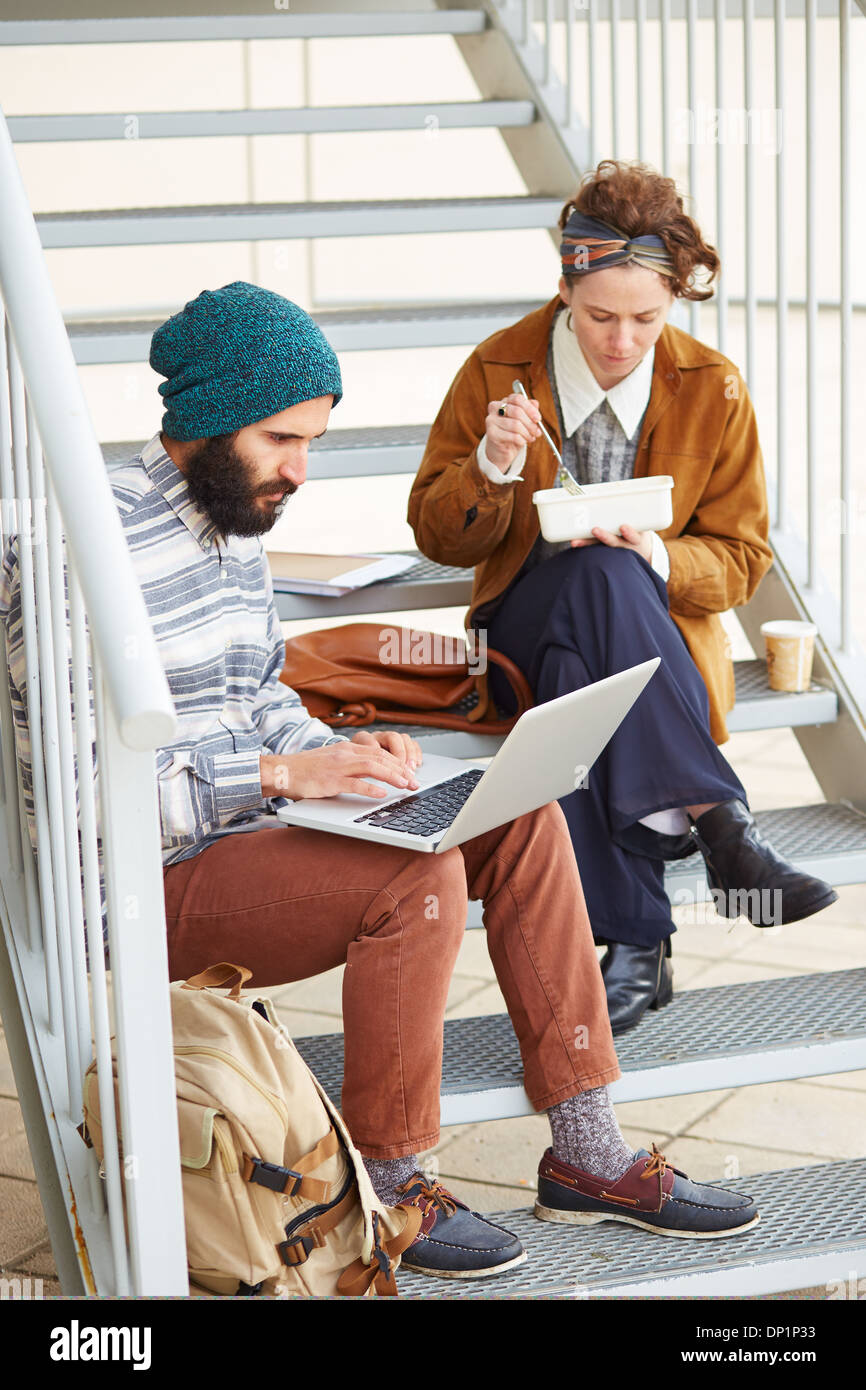 Hipster couple using computer and eating lunch sitting in stairs at university campus Stock Photo