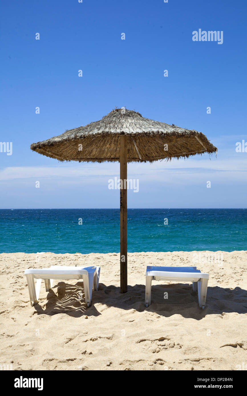 Two sunbathing long chairs and straw parasol on the beach. Stock Photo