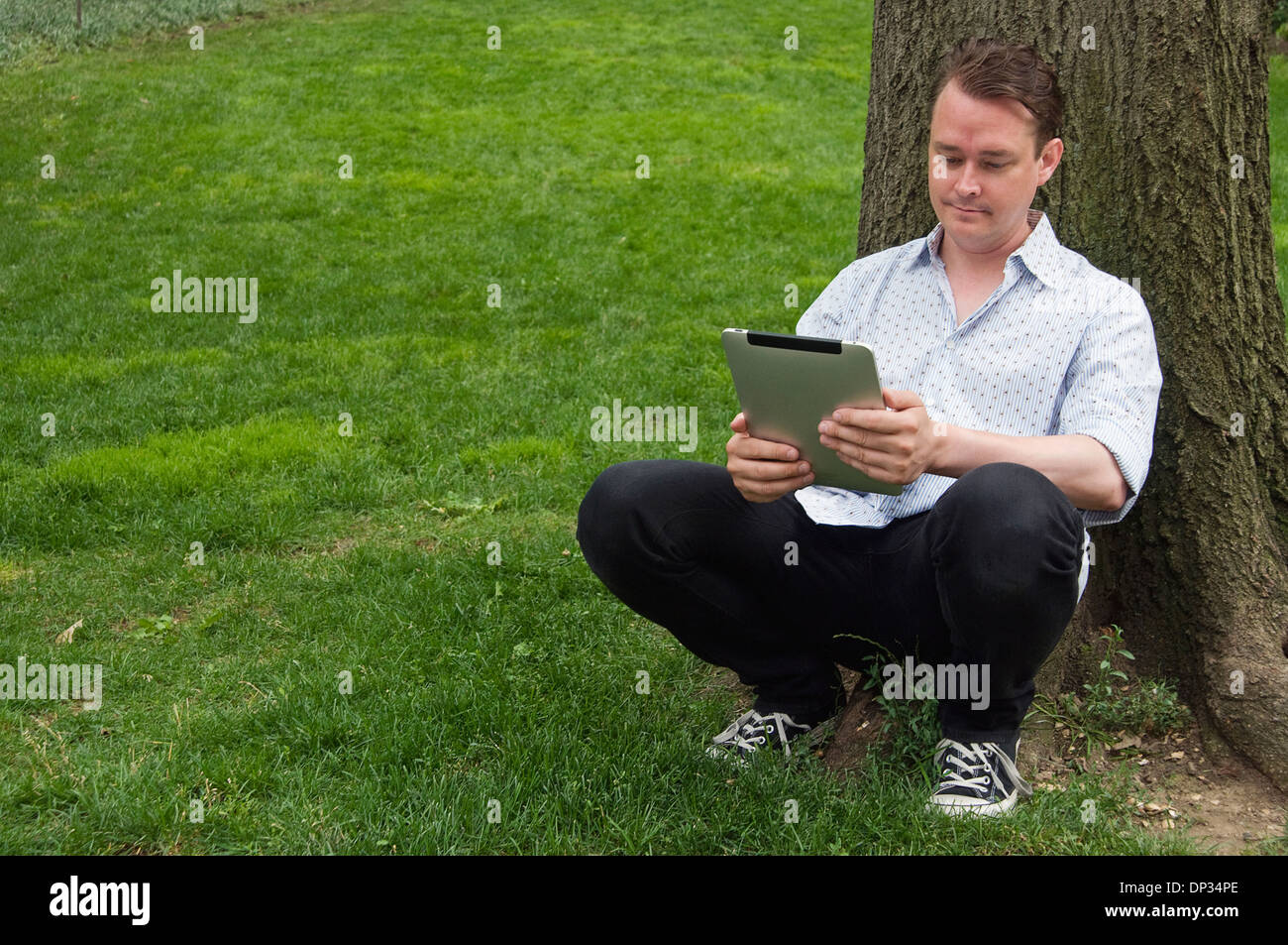 A man leaning against a tree holding an iPad 3g 64gb Stock Photo