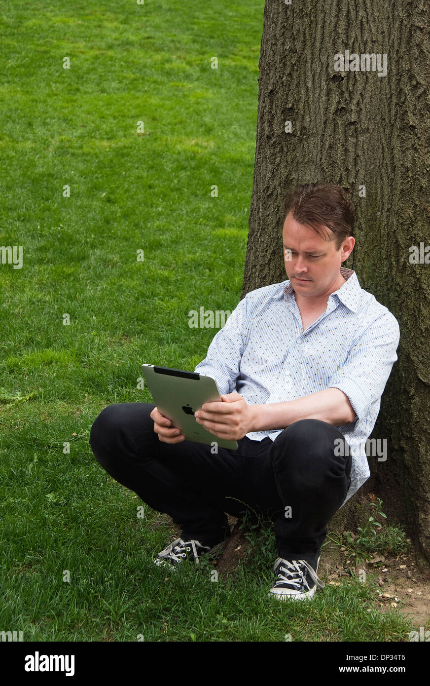 A man leaning against a tree holding an iPad 3g 64gb Stock Photo