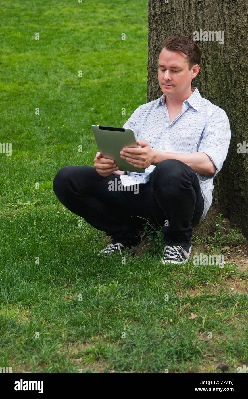 A man leaning against a tree holding an iPad 3g 64gb Stock Photo