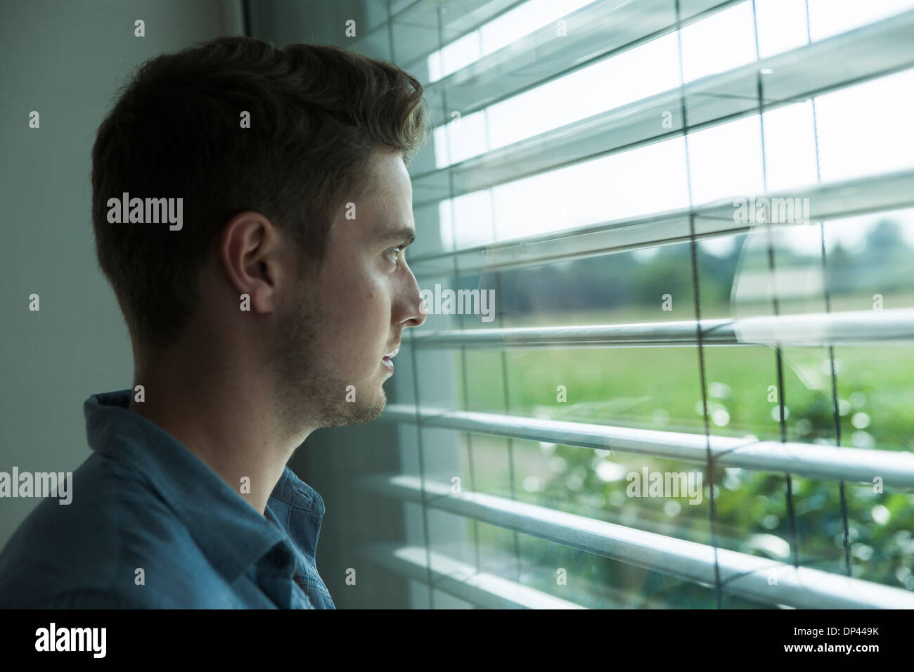 Close-up portrait of young man, looking out window through blinds, Germany Stock Photo