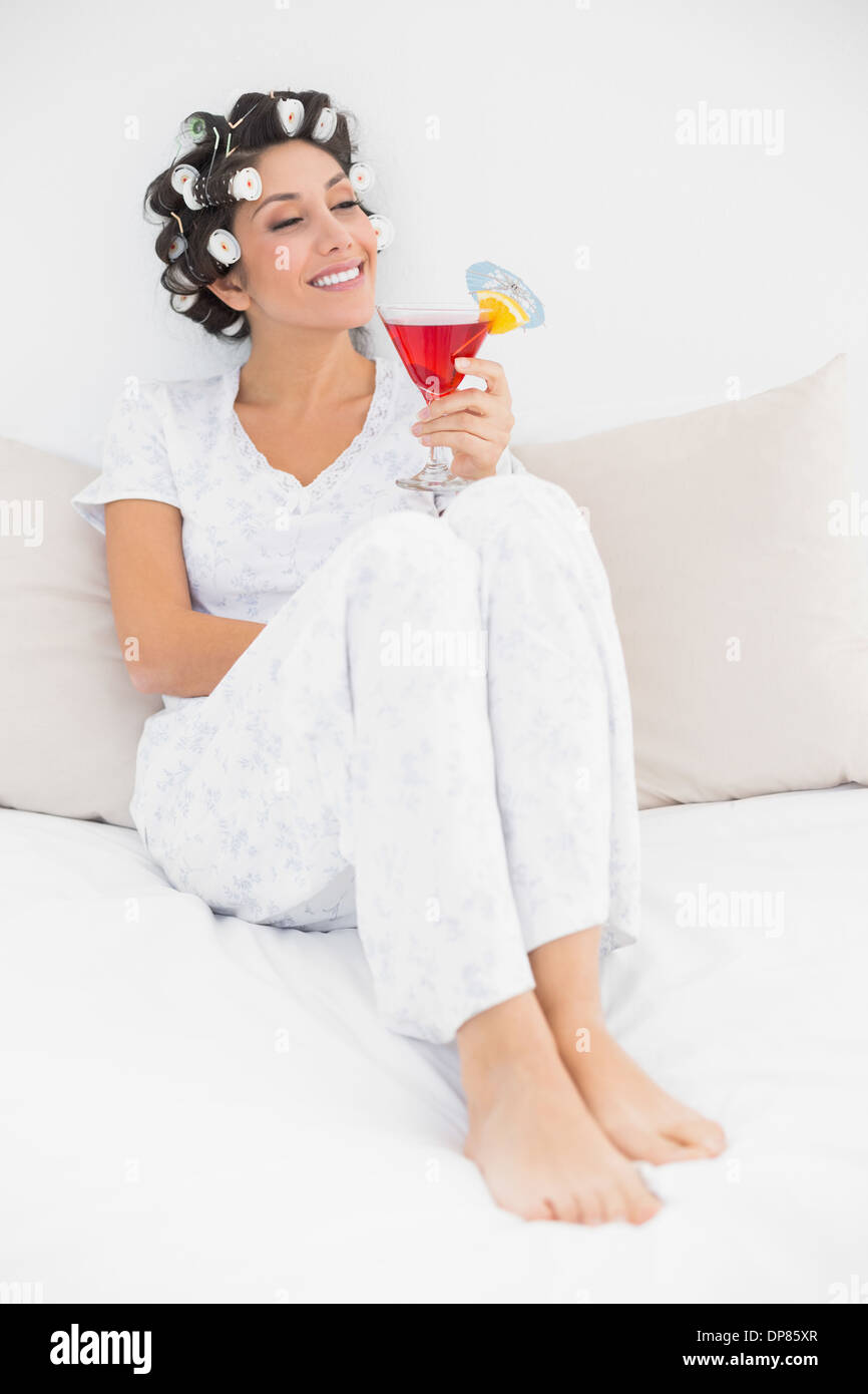 Content brunette in hair curlers sitting on her bed holding a cocktail Stock Photo