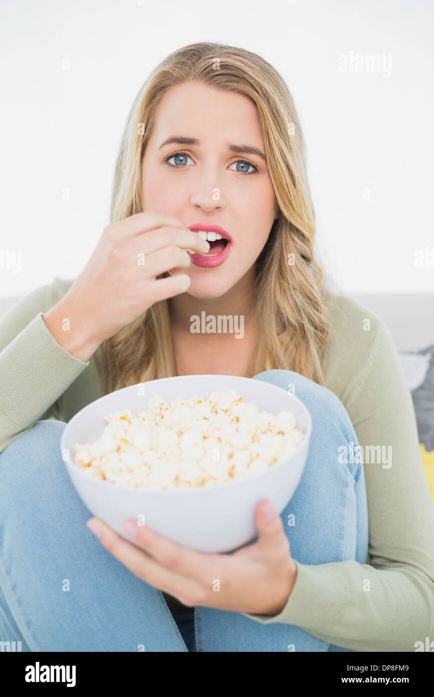 Surprised pretty blonde eating popcorn sitting on cosy sofa Stock Photo