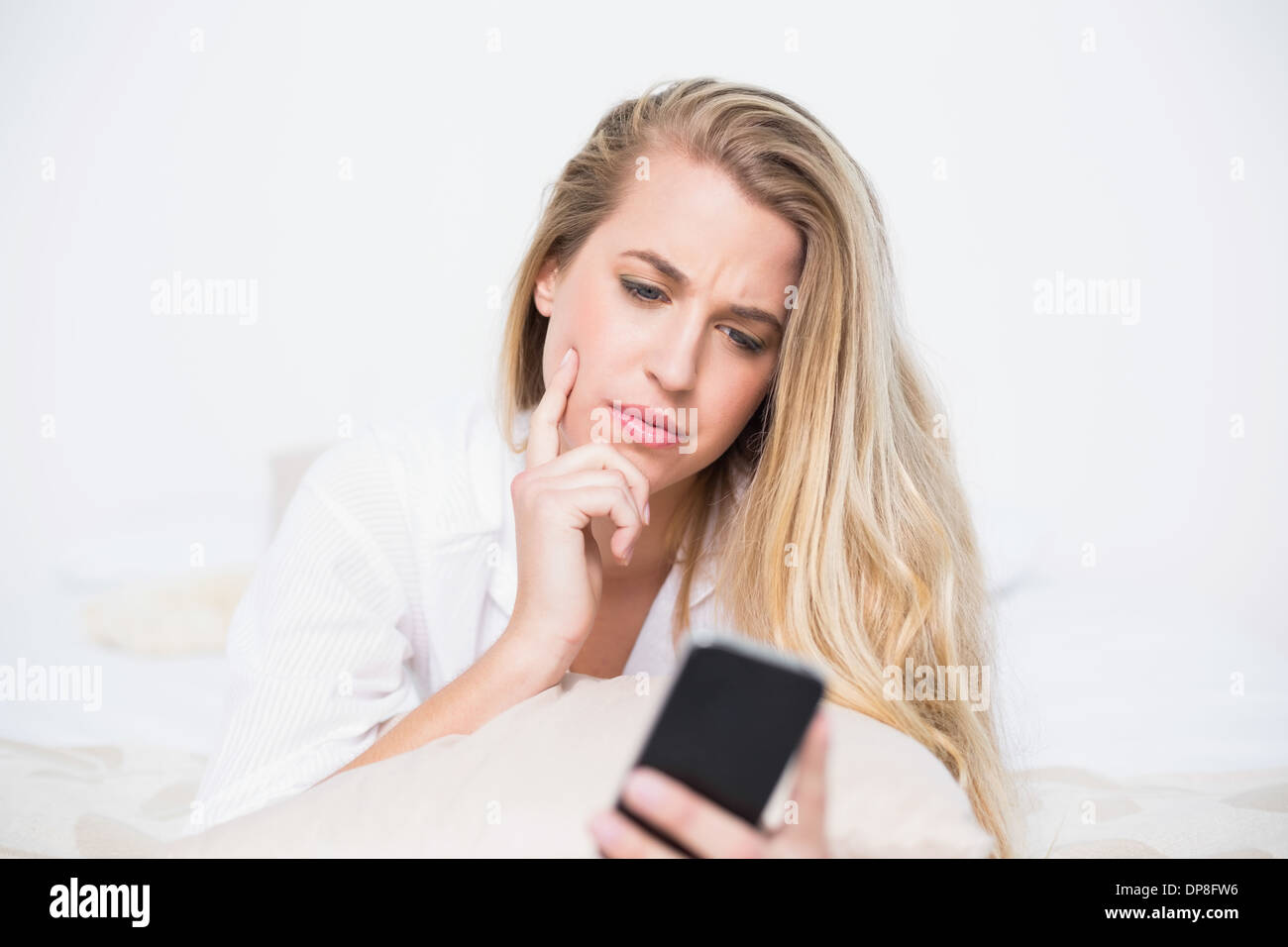 Frowning model holding her smartphone lying on cosy bed Stock Photo
