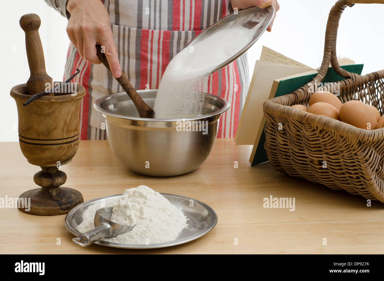 Woman baking a cake, adding the sugar to a bowl & surrounded by the other ingredients. Stock Photo