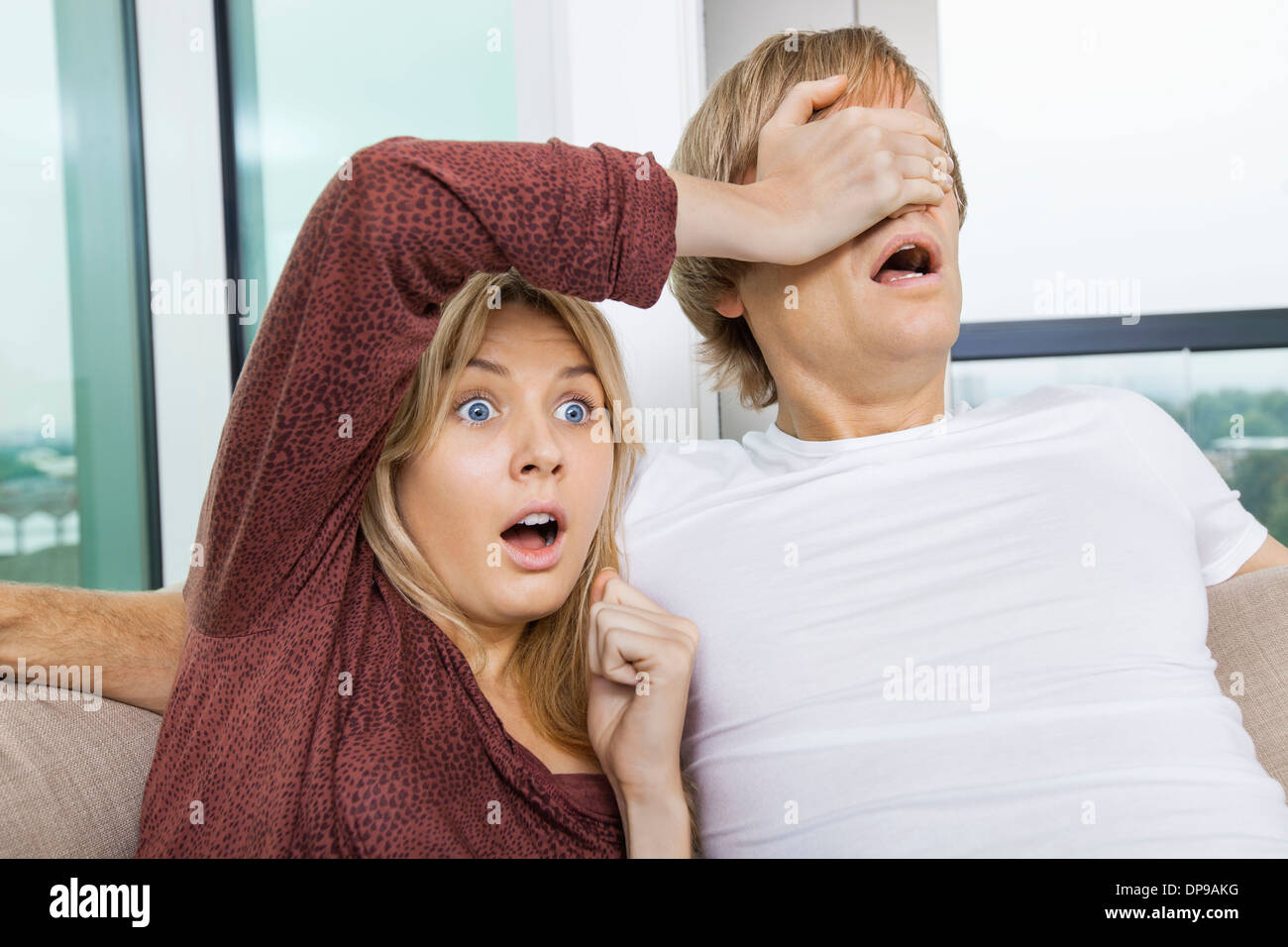 Shocked woman covering man's eyes while watching TV at home Stock Photo