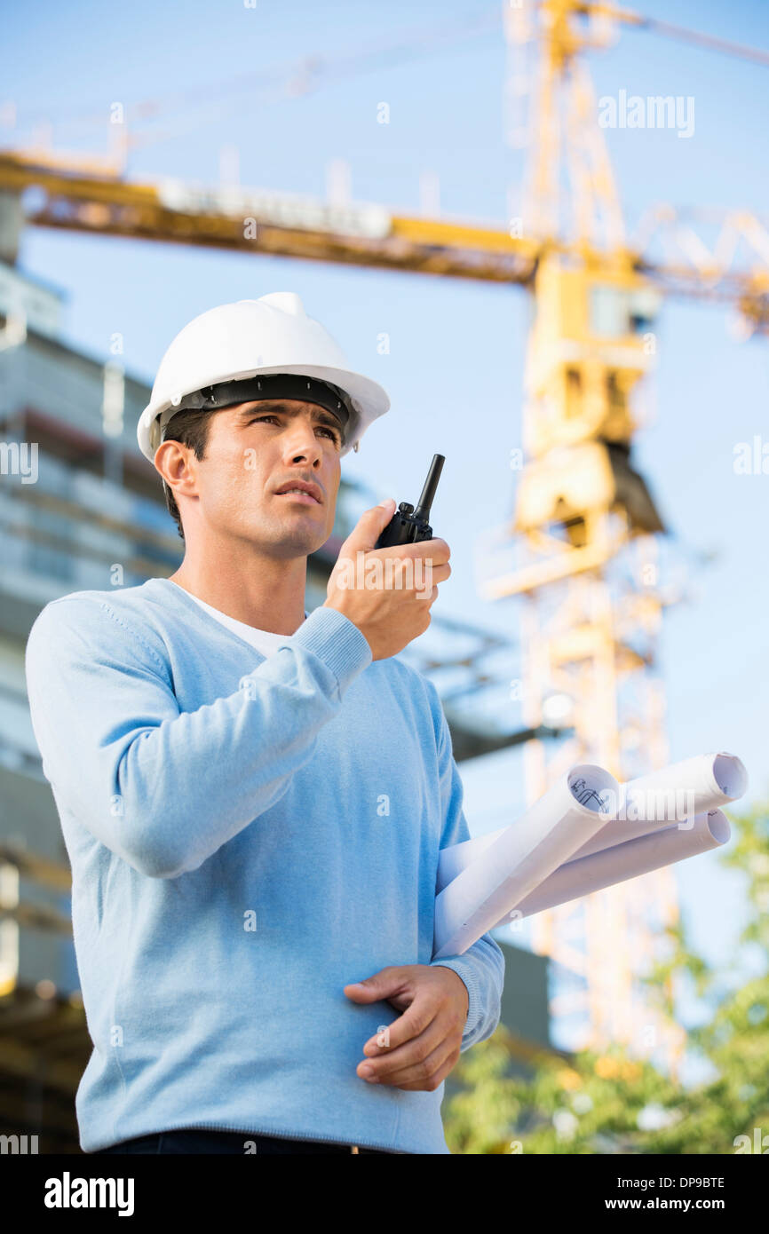 Male architect holding blueprints while using walkie-talkie at construction site Stock Photo