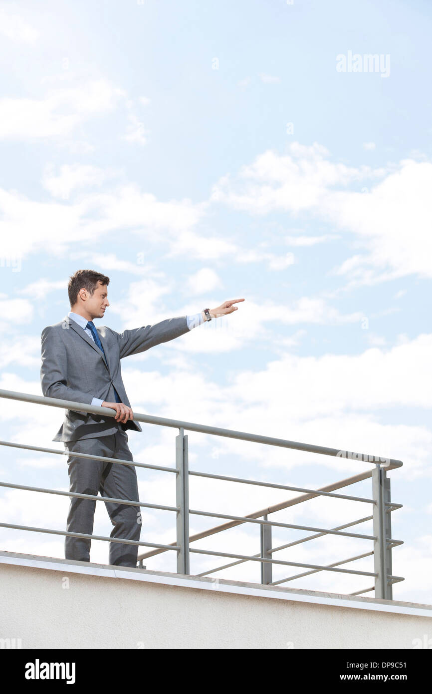 Young businessman with arm raised standing at terrace railings against sky Stock Photo