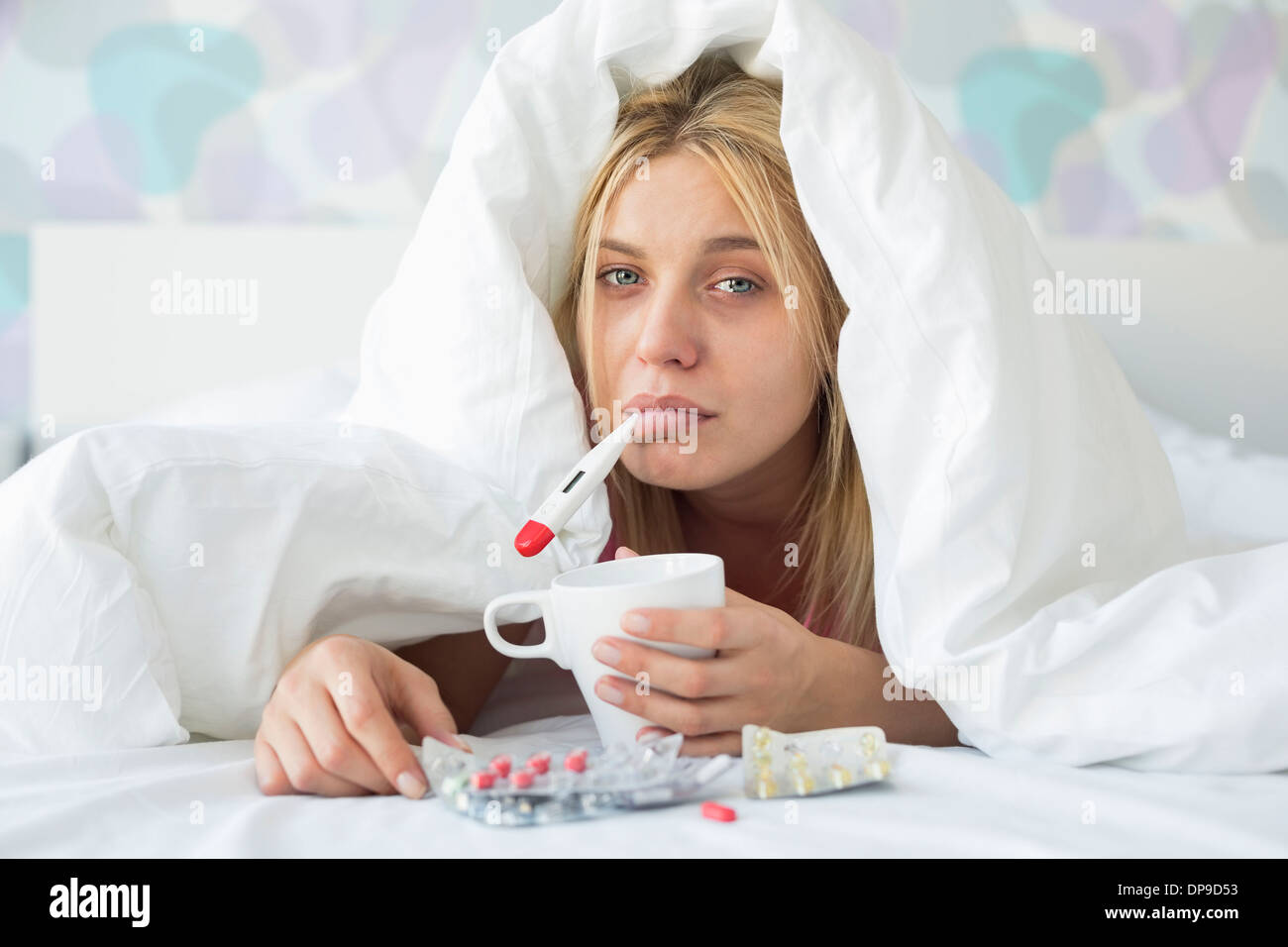 Portrait of sad woman with coffee mug taking temperature while wrapped in quilt on bed Stock Photo