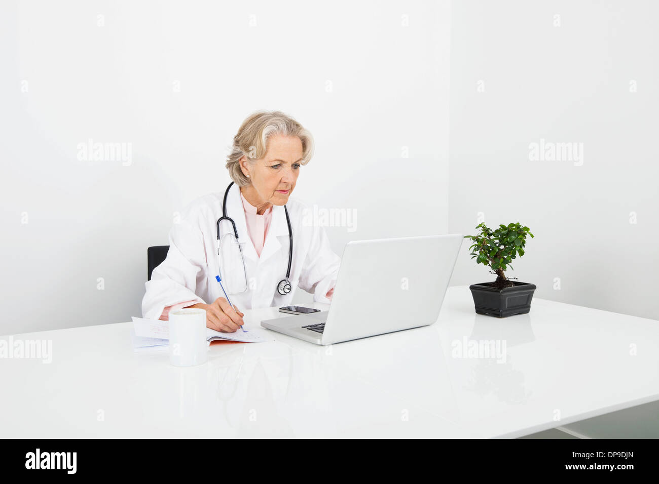 Senior female doctor writing notes while looking at laptop in clinic Stock Photo