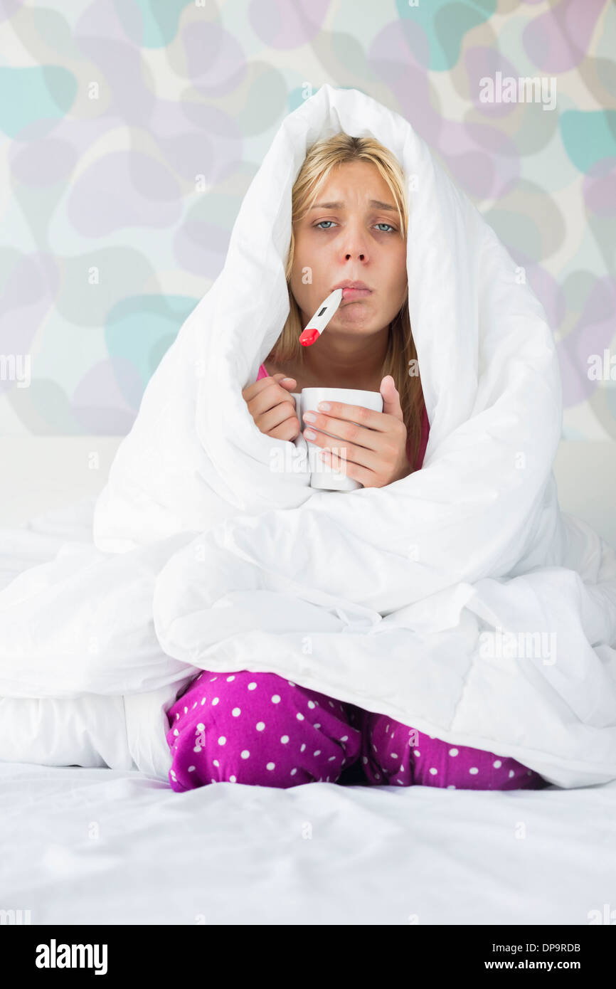 Portrait of young woman with coffee mug taking temperature while wrapped in quilt on bed Stock Photo