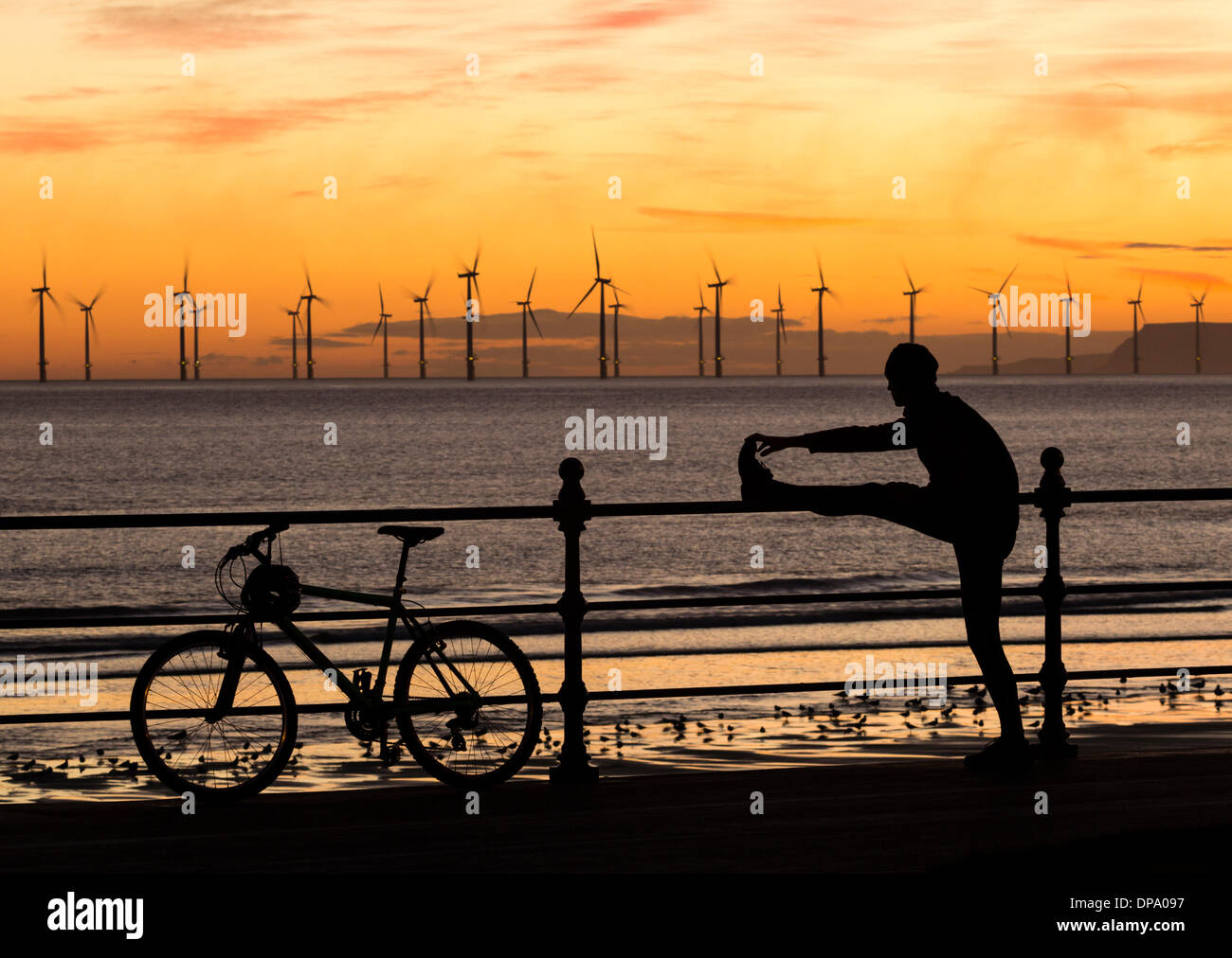 Mountain biker stretching at sunrise on Seaton Carew beach with Teesside Offshore Wind farm in background. UK Stock Photo
