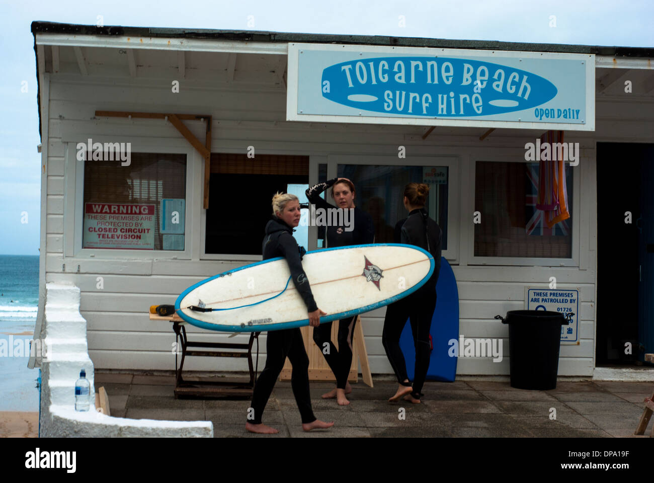 Female surfers in wetsuits with surf board outside a surf hire kiosk at Toicarne Beach in Cornwall Stock Photo