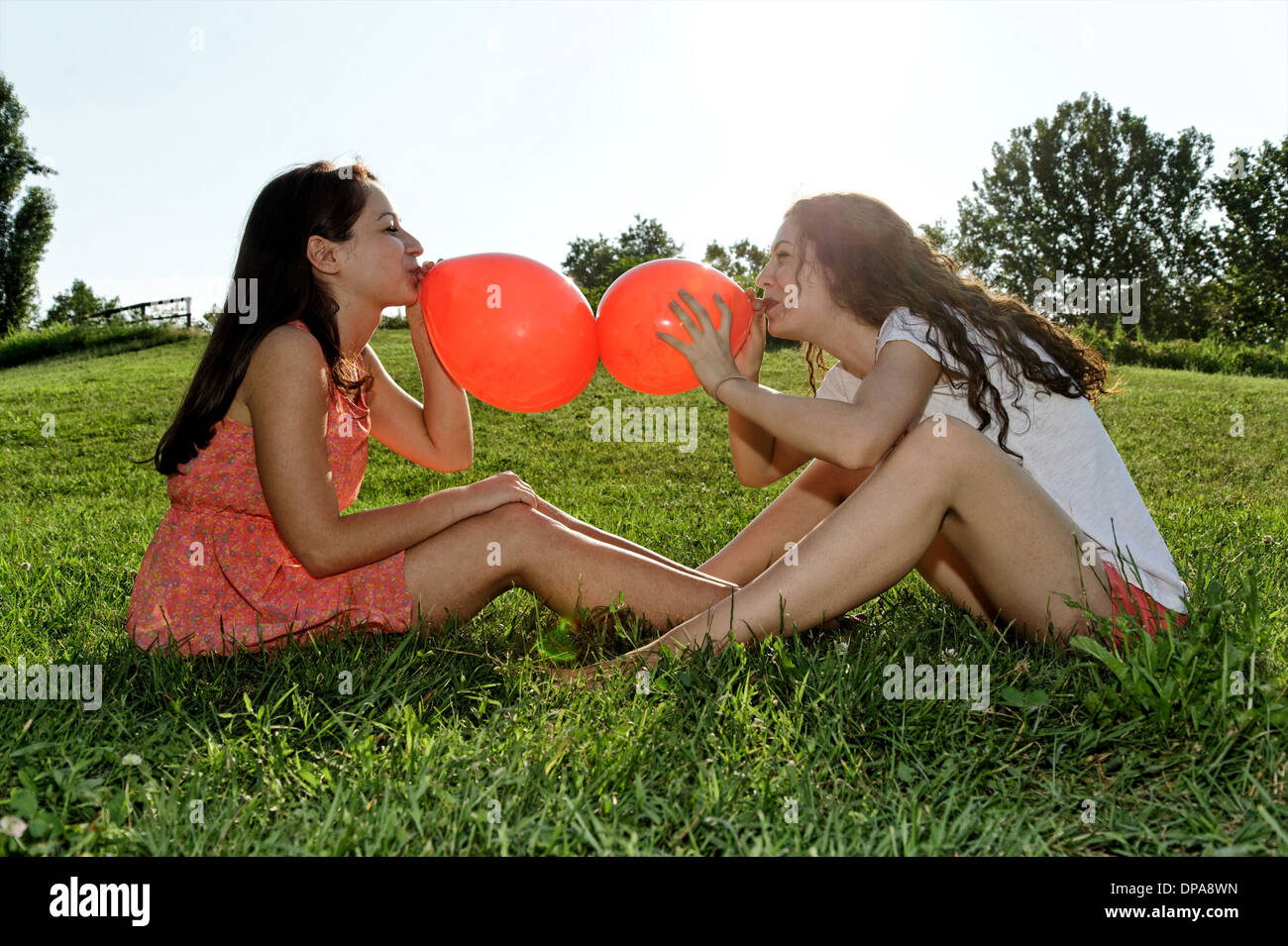 Two young women sitting on grass bowing up red balloons Stock Photo