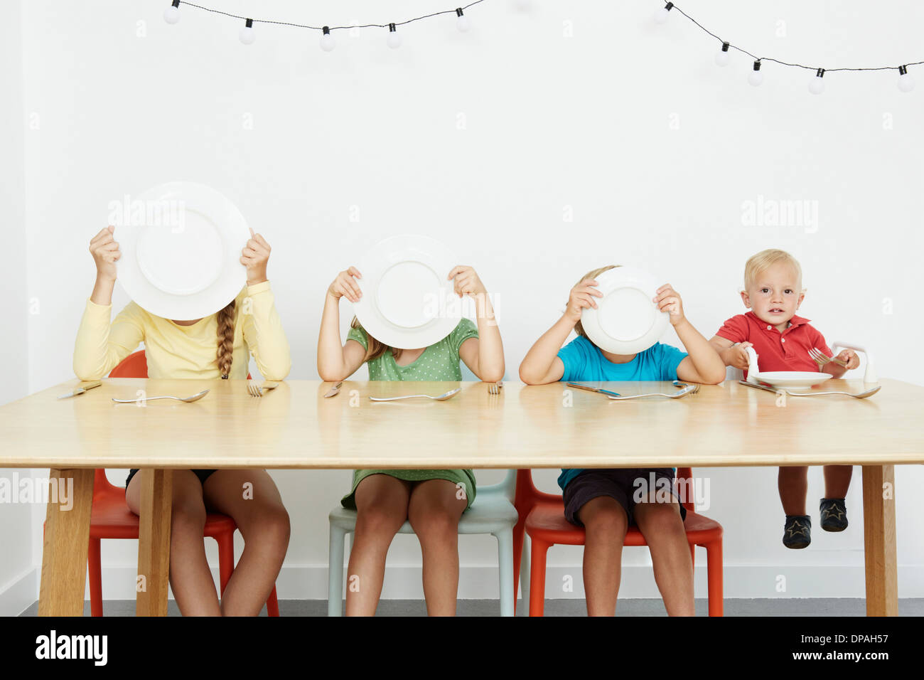 Four children sitting at table, three covering faces with plates Stock Photo