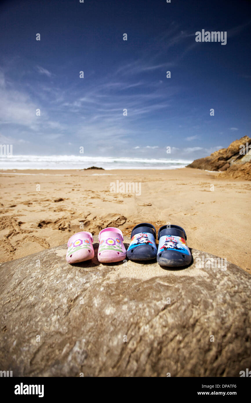 Childrens 'crocs' shoes on a rock on a beach with waves and blue skies behind. Stock Photo