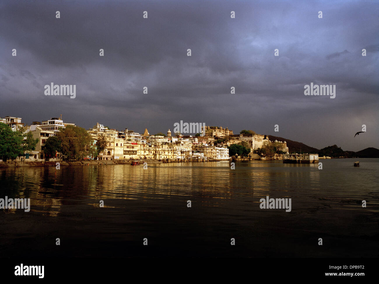 A view across Lake Pichola to the bathing ghats and the City Palace in Udaipur in Rajasthan in India in South Asia. Sunset Sky Stormy Weather View Stock Photo