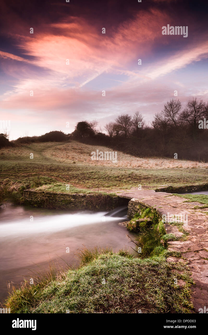 The sun rises behind the little stone Clapper bridge at Daniels Well in the Wiltshire hillside town of Malmesbury. Stock Photo