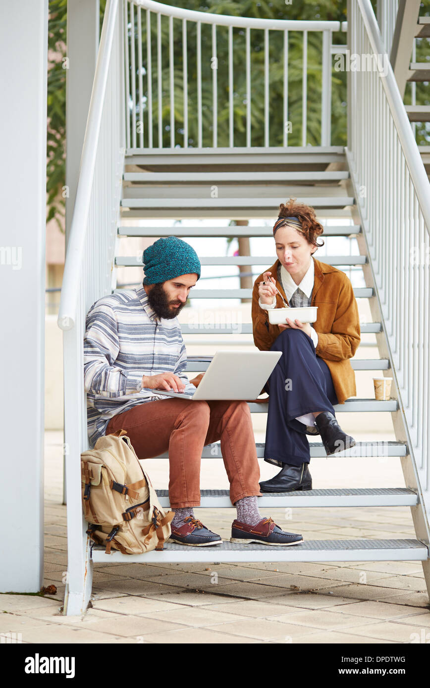 Hipster couple using computer and eating lunch sitting in stairs at university campus Stock Photo