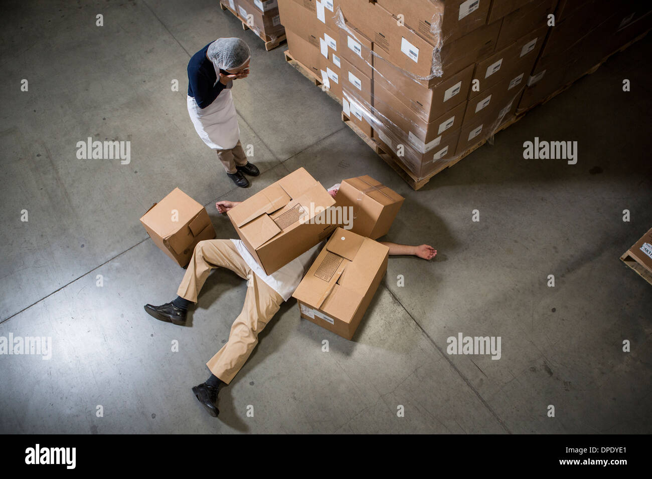 Woman looking at man lying on floor covered by cardboard boxes in warehouse Stock Photo
