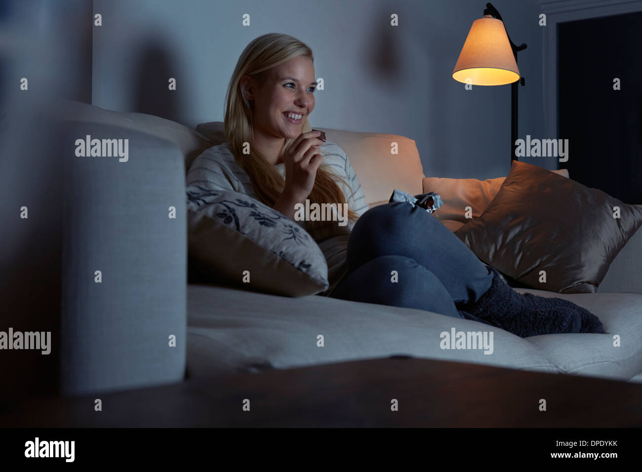 Young woman sitting on sofa, watching tv, eating chocolate Stock Photo