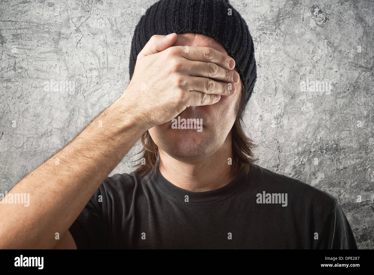 Portrait of Casual Man with Black Cap covering face in disbelief against grunge background Stock Photo
