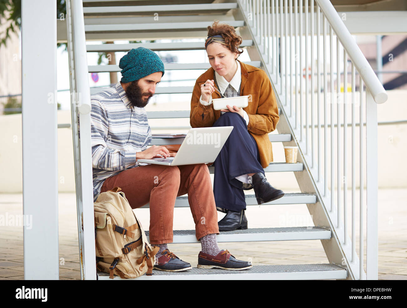 Hipster couple using computer and eating lunch sitting in stairs at university campus Stock Photo