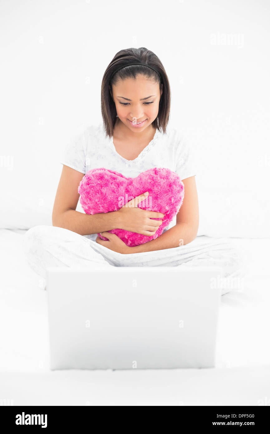 Concentrated young dark haired model cuddling a pillow and looking at a laptop Stock Photo