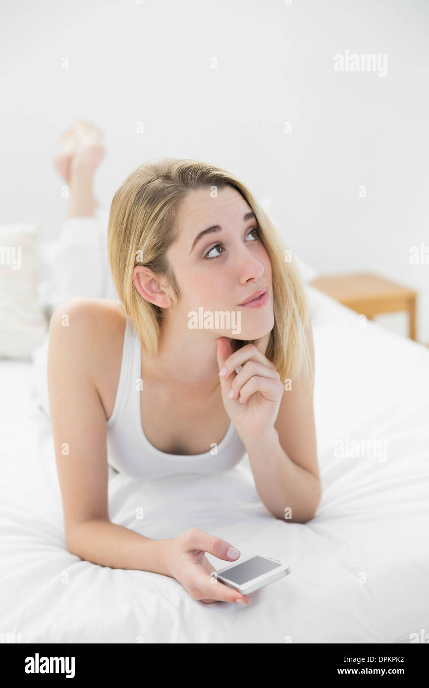 Thoughtful blonde woman holding her smartphone lying on her bed Stock Photo