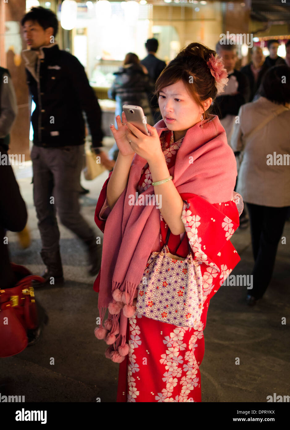 Young Japanese girl wearing a flowery red winter kimono on a cool autumn evening, while using a smartphone. Stock Photo