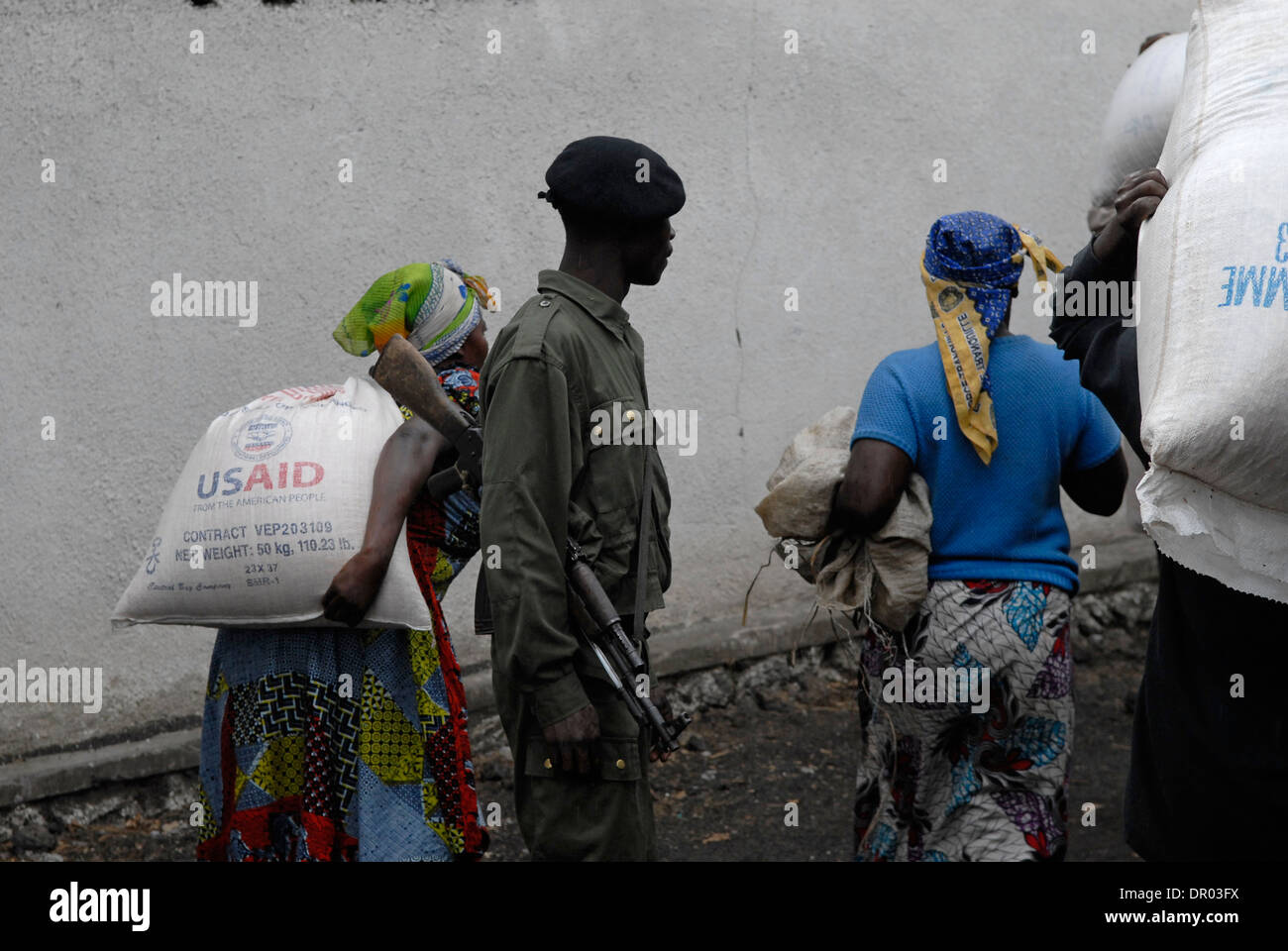 An armed FARDC Congolese government soldier stands guard as internally displaced women carry large sack of maize donated by USAID agency during food distribution carried out by the World Food Programme WFP in North Kivu province, Congo DR Africa Stock Photo