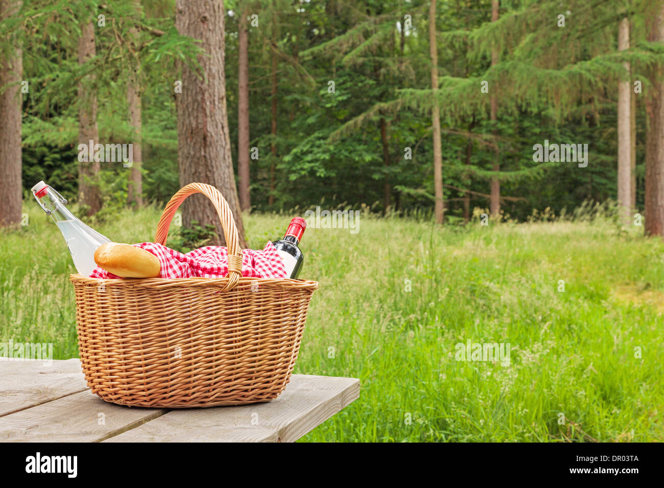 A whicker picnic basket full of food and drink on a table in a woodland setting on a bright summers day. Stock Photo