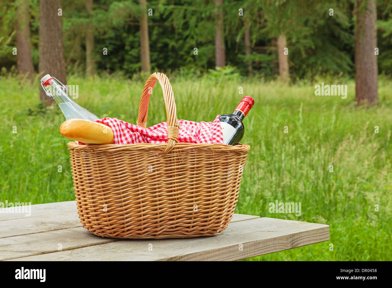A whicker picnic basket full of food and drink on a table in a woodland setting on a bright summers day. Stock Photo