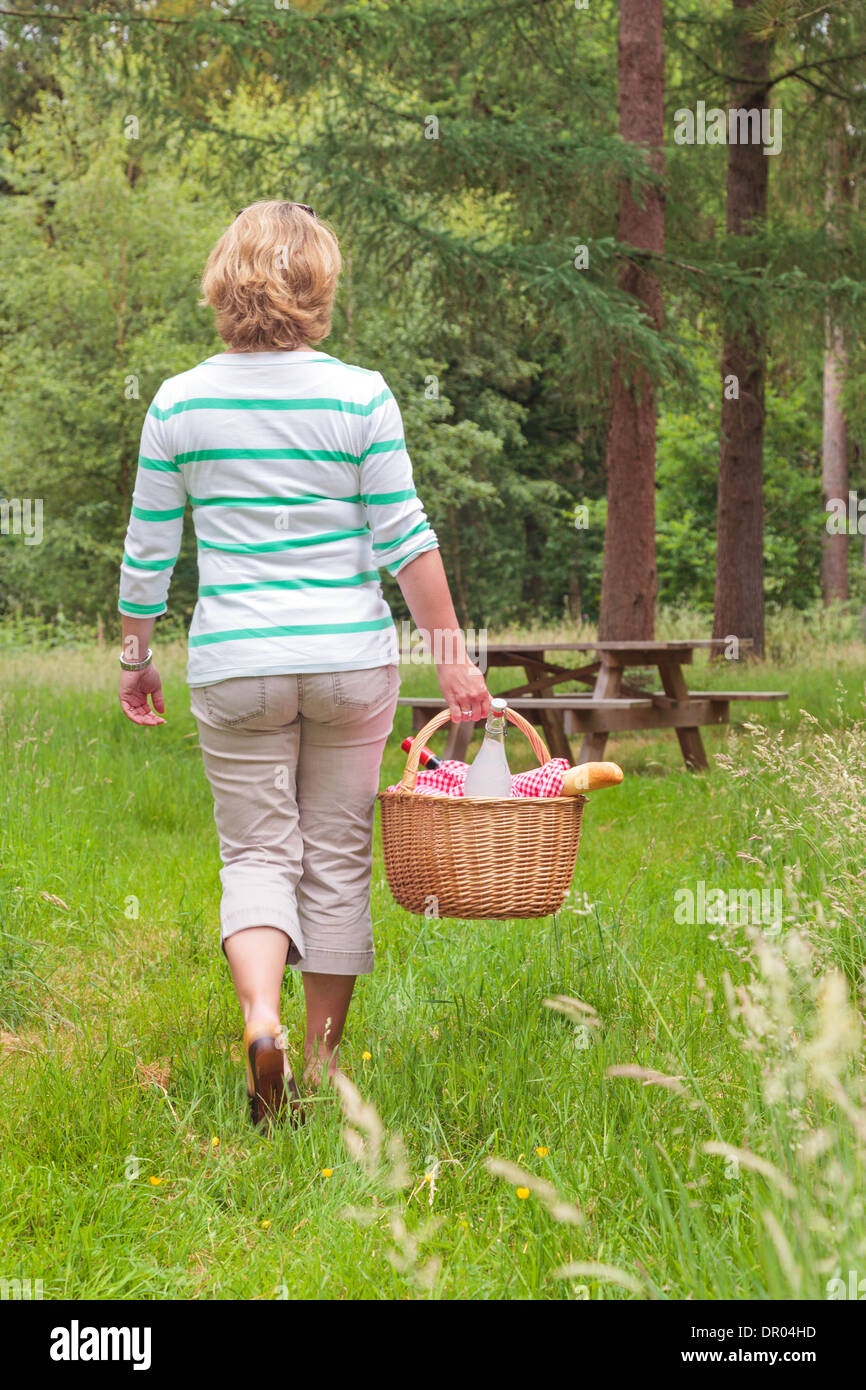 A woman carrying a whicker picnic basket full of food and drink towards a table in woodland, on a bright summers day. Stock Photo