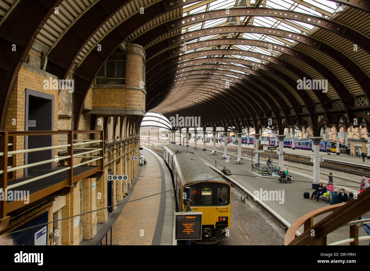 Interior view of trainshed with iron & glass roof, stationary trains & people waiting on platfom - York Railway Station, North Yorkshire, England, UK. Stock Photo
