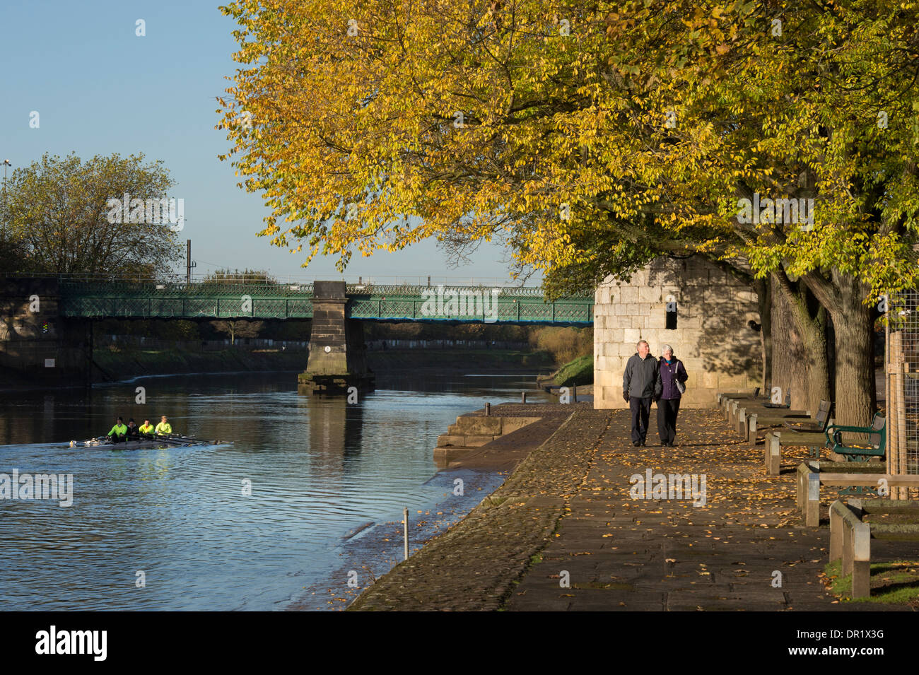 Rowers in boat & couple walking - quiet, scenic, sunlit, tree-lined riverside footpath in autumn - River Ouse, Dame Judi Dench Walk, York, England, UK Stock Photo