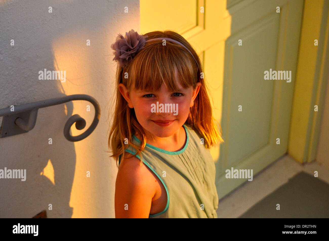 portrait of little blonde girl smiling on holiday looking up at the camera as she is bathed in sunlight in Santorini Greece Stock Photo