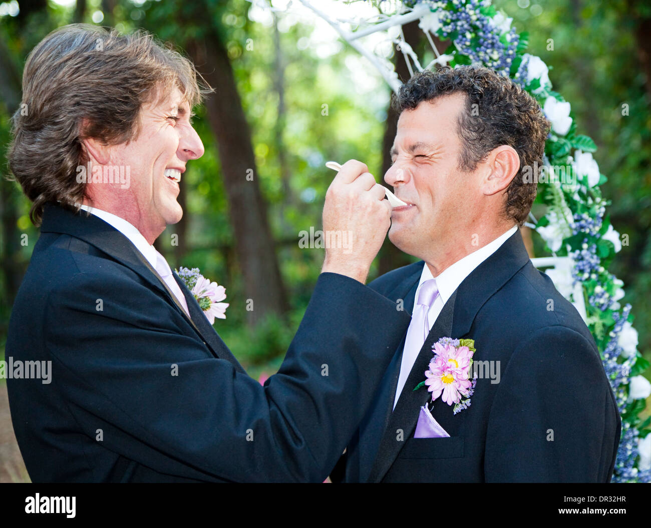 One groom at a gay wedding feeding cake to his husband and laughing.  Stock Photo